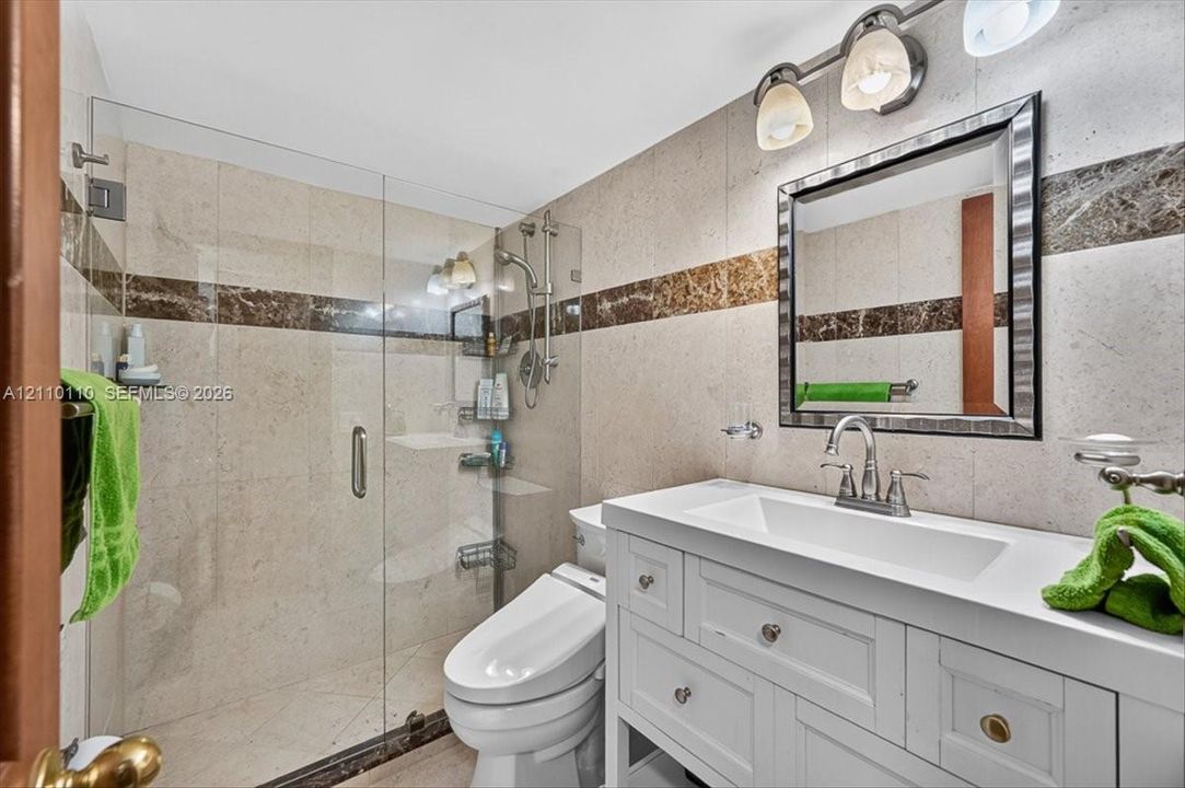 White vanity with integrated rectangular basin and metal hardware, walk-in glass-enclosed shower, stone wall tiling with decorative inlay, and wall-mounted vanity lighting