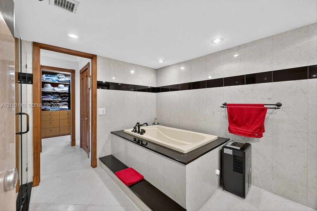 Soaking tub featuring a black stone surround and deck, recessed lighting, floor-to-ceiling wall tile with black accent inlay, and wood-finish built-in closet cabinetry