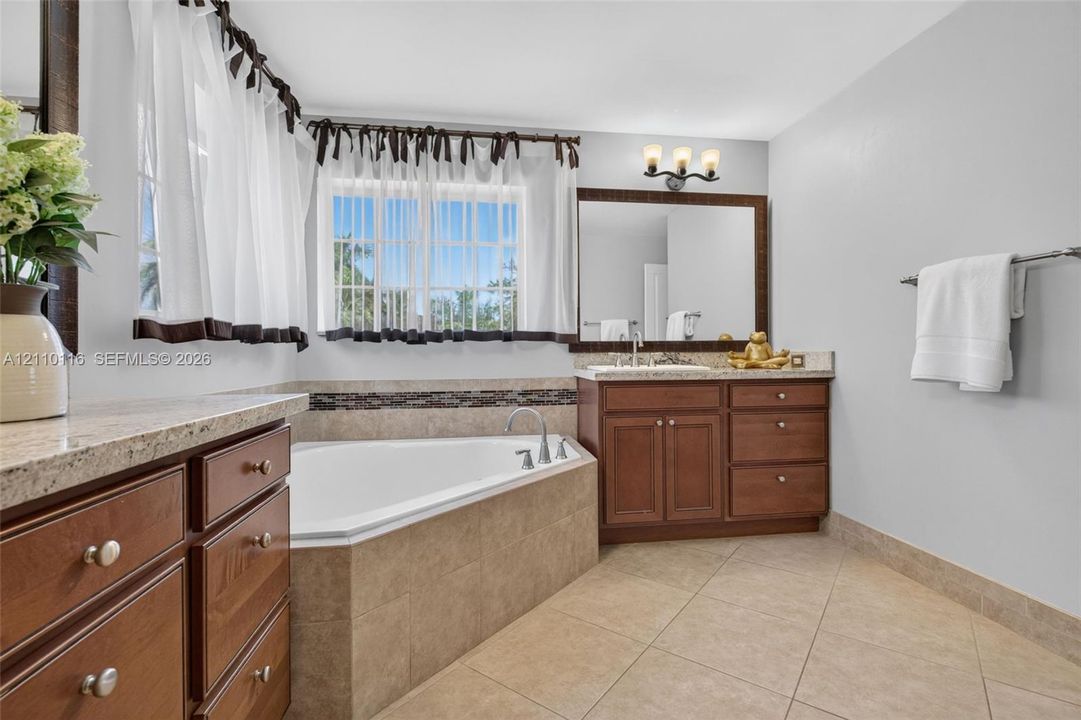 Bathroom featuring a corner soaking tub with tile surround, wood cabinetry, stone-finish countertops, ceramic tile flooring, and a large framed vanity mirror