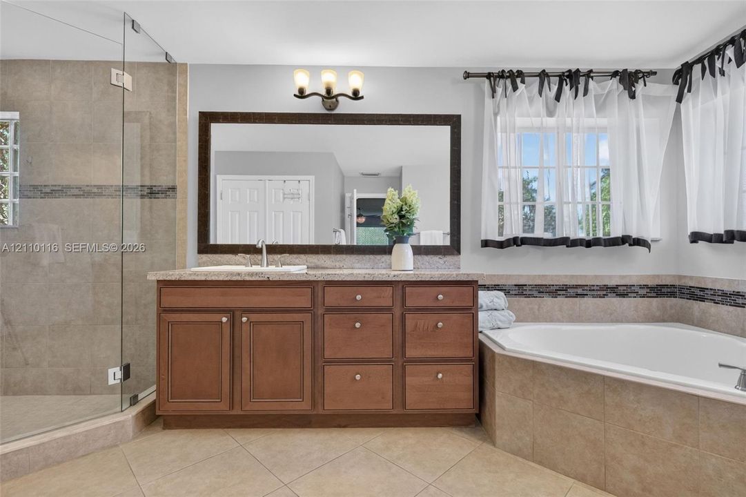 Bathroom featuring wood-finish cabinetry, stone countertop, corner soaking tub with tile surround, glass-enclosed shower, and large framed wall mirror