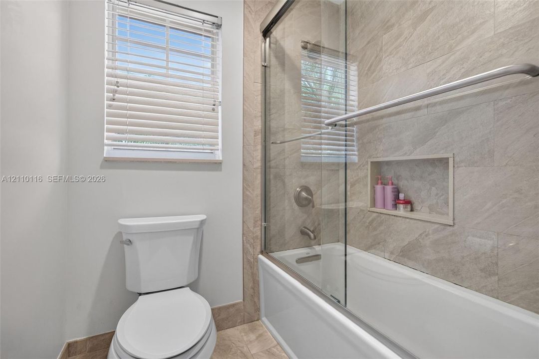 Bathroom featuring a sliding glass shower door, recessed wall niche, neutral stone-tile surround, and window with blinds