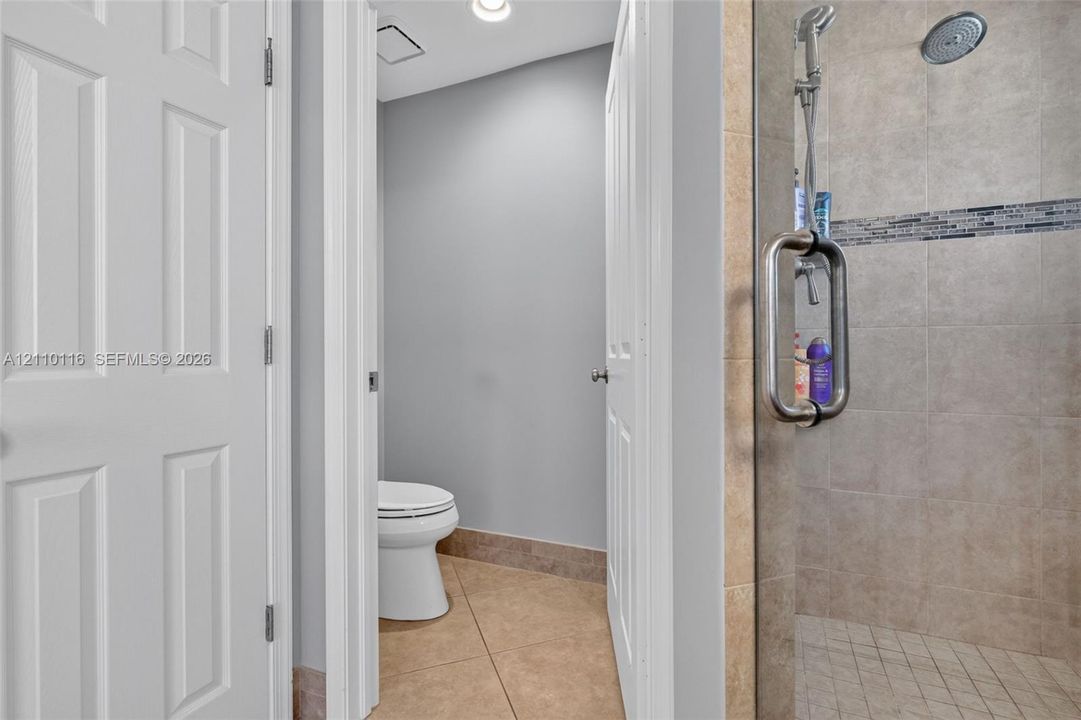 Walk-in shower with a glass door, rainfall showerhead, and tile surround featuring a decorative mosaic accent band, accompanied by tile flooring and white panel doors
