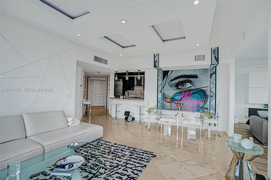 Open-concept living space featuring stone-finish flooring, recessed ceiling lighting with geometric coffers, a pass-through kitchen bar, and a wall accent with integrated lighting