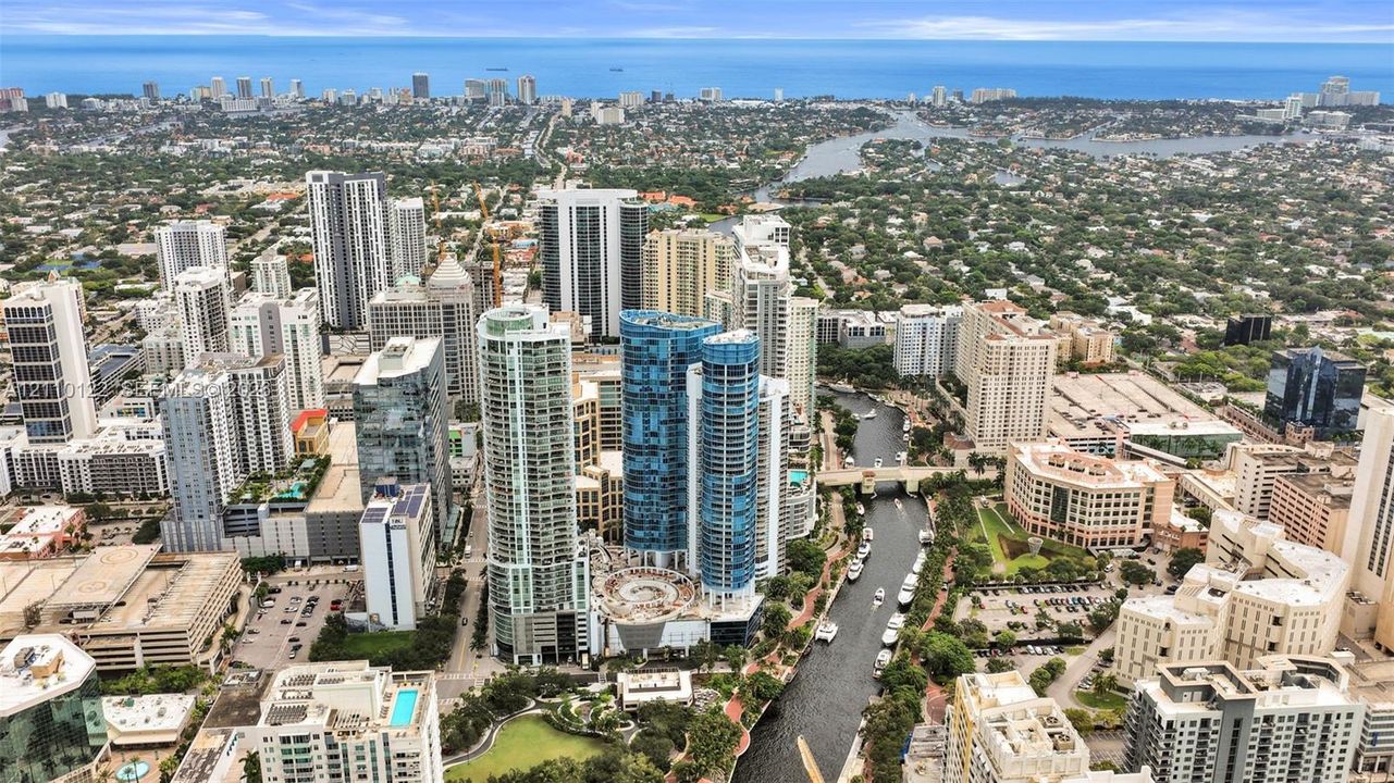 Aerial perspective of urban high-rise development featuring glass-facade towers, proximity to waterway, surrounding metropolitan infrastructure, and coastal horizon