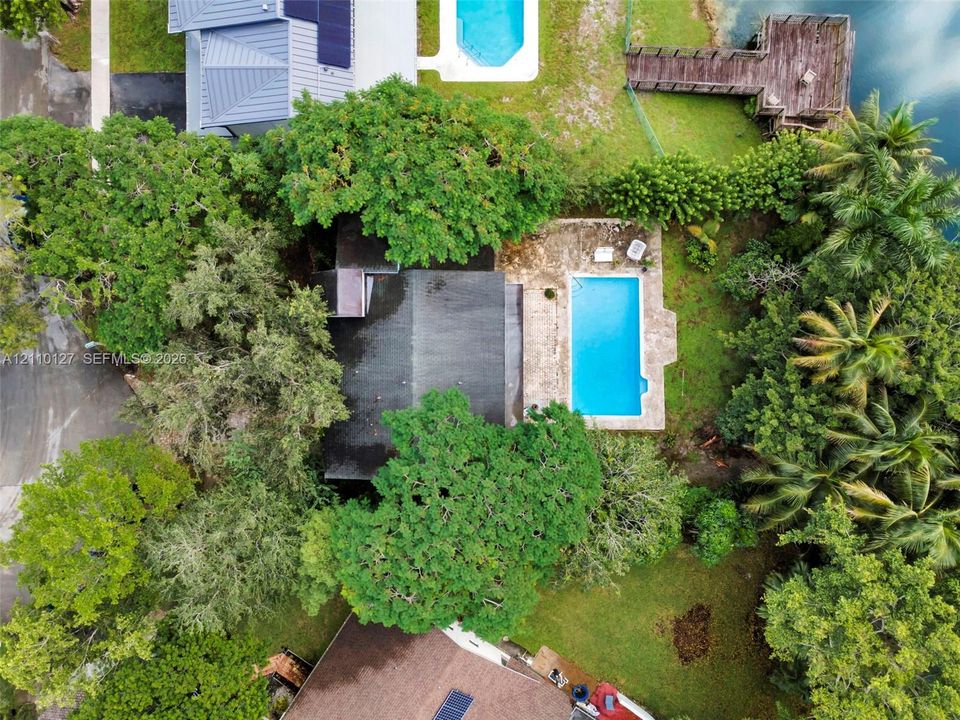 Aerial perspective featuring a private swimming pool, stone patio, wooden dock, and mature canopy trees on a waterfront lot