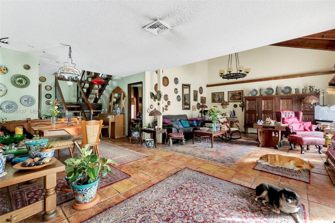 Open-concept living space featuring vaulted ceilings with exposed wood beams, terracotta tile flooring, a custom built-in dark wood cabinetry unit, and an open-tread staircase with wood handrails