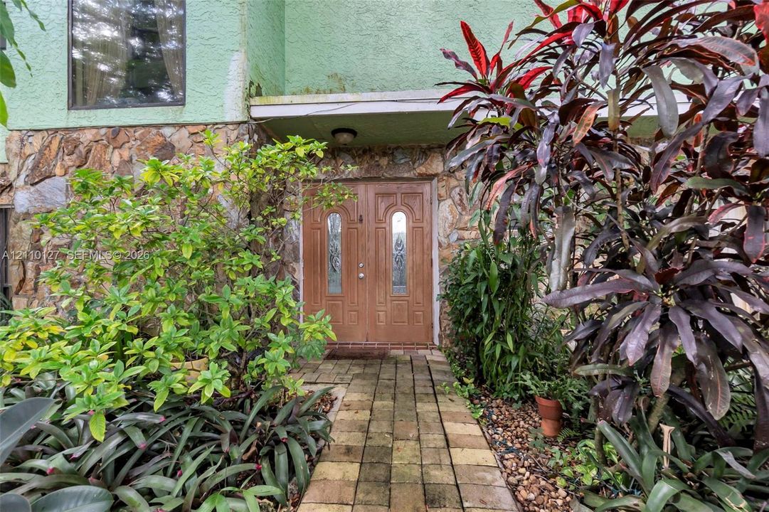 Exterior entrance featuring double doors with decorative glass inserts, natural stone masonry facade, stucco detailing, and a brick paver walkway