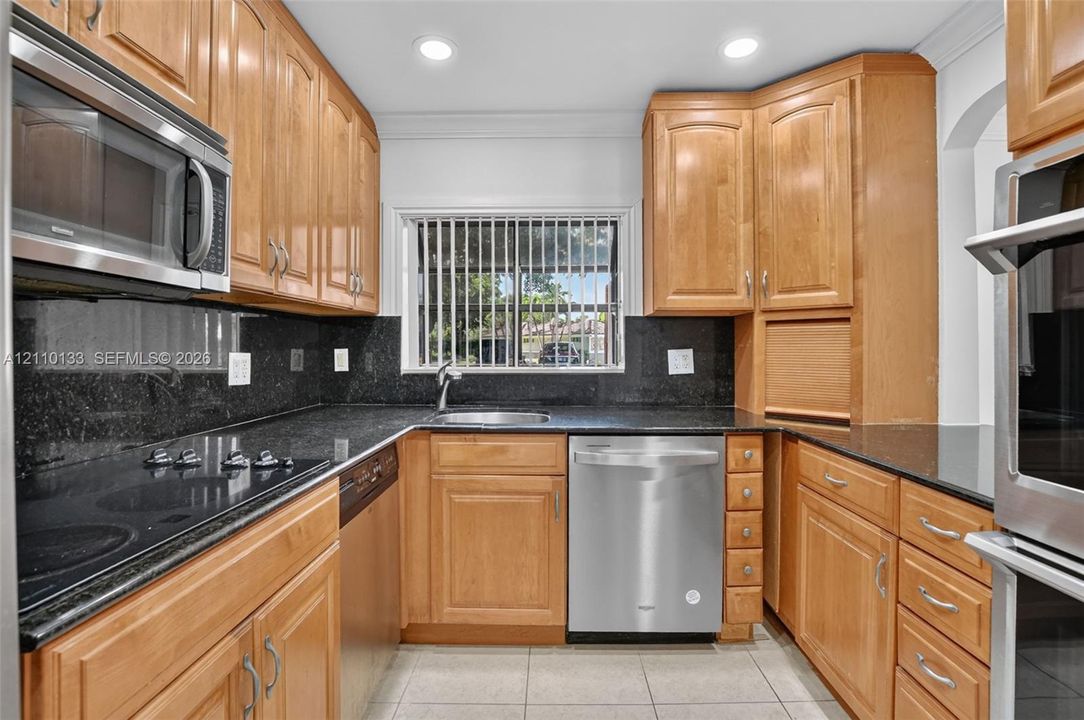 Kitchen featuring warm-tone wood cabinetry, dark stone-finish countertops, stainless steel appliances, recessed ceiling lighting, and tile flooring