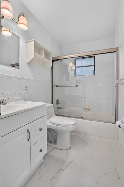 Bathroom featuring a white vanity with integrated sink, stone-patterned tile flooring, three-light wall-mounted fixture, and a glass-enclosed tub with shower surround