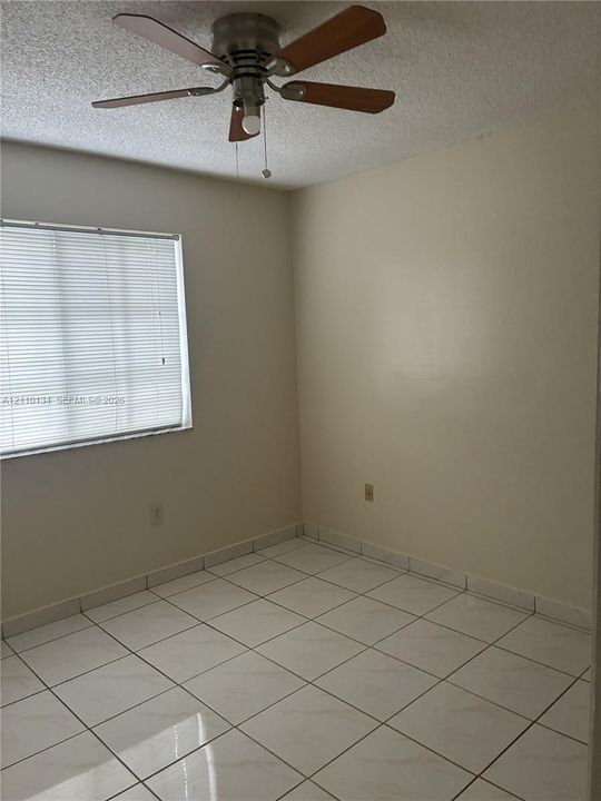 Room featuring light-colored tile flooring, neutral wall tones, a ceiling fan with four wooden-finish blades, and a window with horizontal blinds