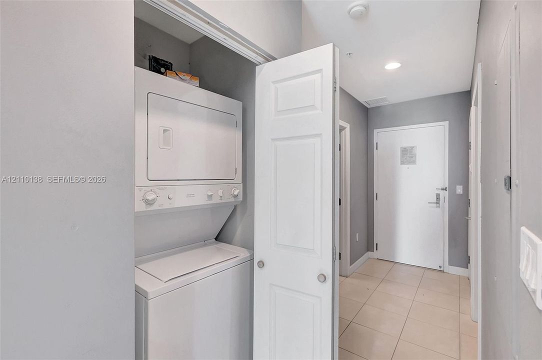 Laundry closet featuring stacked white appliance set, recessed ceiling lighting, and neutral-toned floor tiling