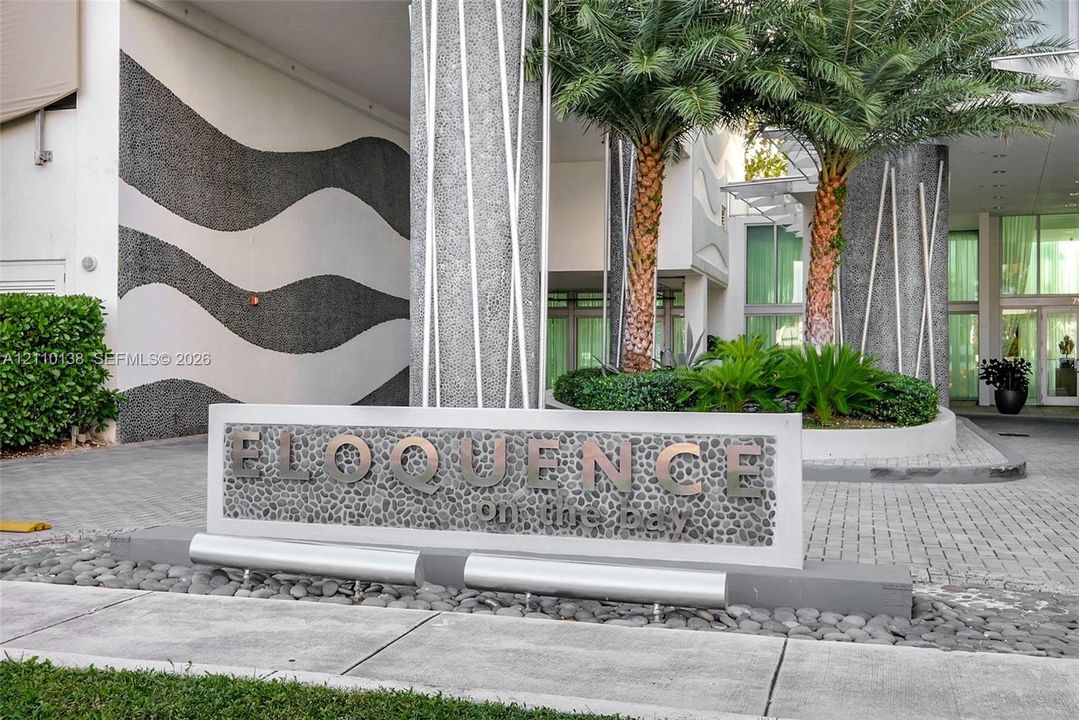 Exterior entry featuring a textured stone-inlay monument sign, paved driveway, sculptural concrete columns, and integrated professional landscaping