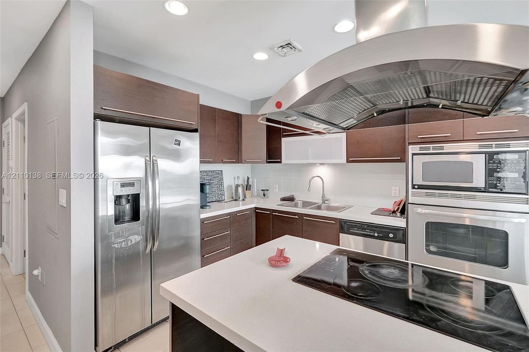 Kitchen featuring dark wood-finish cabinetry, solid surface countertops, stainless steel appliances, a built-in oven and microwave, and a stainless steel range hood