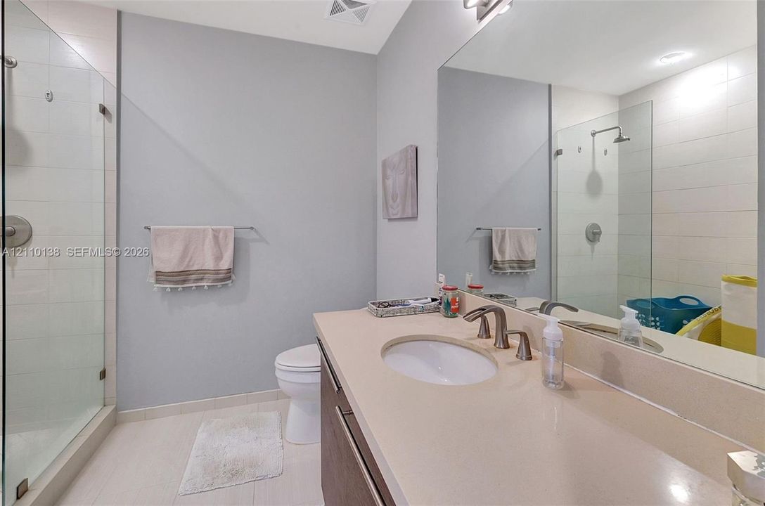 Bathroom featuring a vanity with a solid-surface countertop, an undermount sink, a glass-enclosed shower with tiled surround, and a large wall-mounted mirror