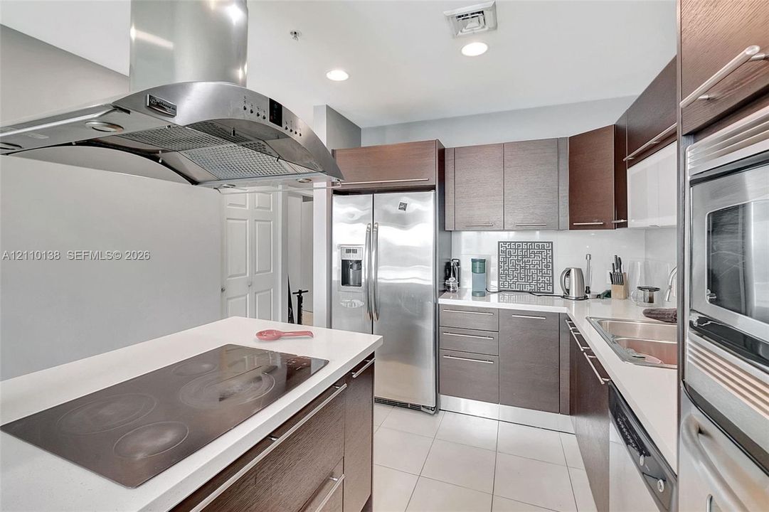 Kitchen featuring custom flat-panel cabinetry, light-colored stone countertops, stainless steel appliances, a ceiling-mounted range hood, and tile flooring