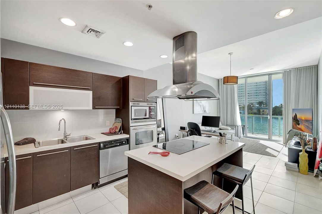 Kitchen featuring dark wood-finish cabinetry, a central island with a cooktop, built-in stainless steel oven and microwave, recessed lighting, and large glass doors