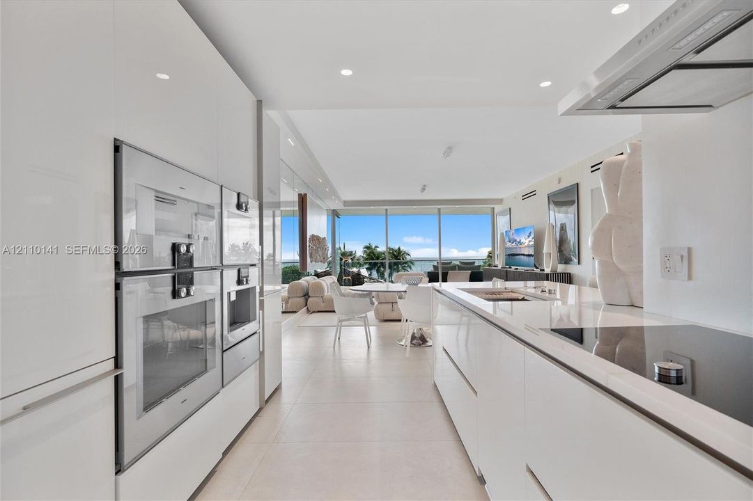 Contemporary kitchen featuring white high-gloss cabinetry, stainless steel built-in double wall ovens, a seamless solid-surface island countertop, integrated cooktop, and floor-to-ceiling glass paneling