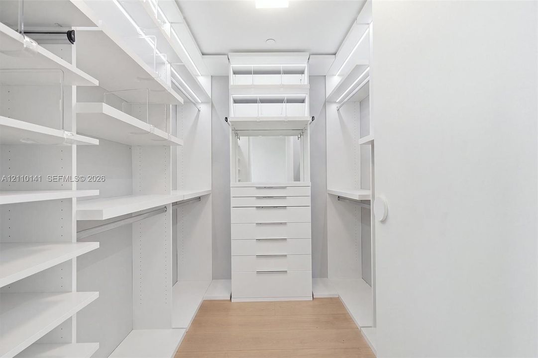 Custom walk-in closet featuring floor-to-ceiling white shelving, integrated LED accent lighting, a center stack of drawers, and wood-finish flooring