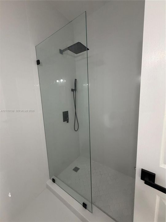Walk-in shower featuring a frameless glass panel, matte black wall-mounted fixtures, a square rain-style showerhead, and a hand-held sprayer