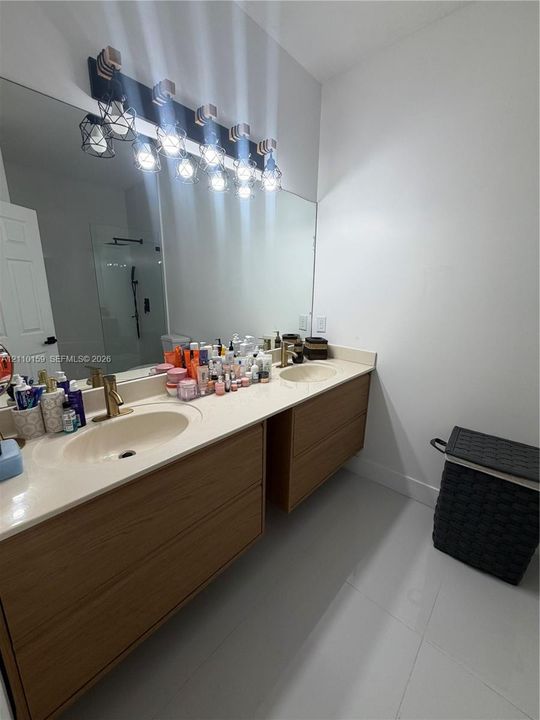 Floating wood-finish vanity with dual integrated sinks, neutral solid-surface countertop, brass-toned fixtures, and a multi-bulb vanity light