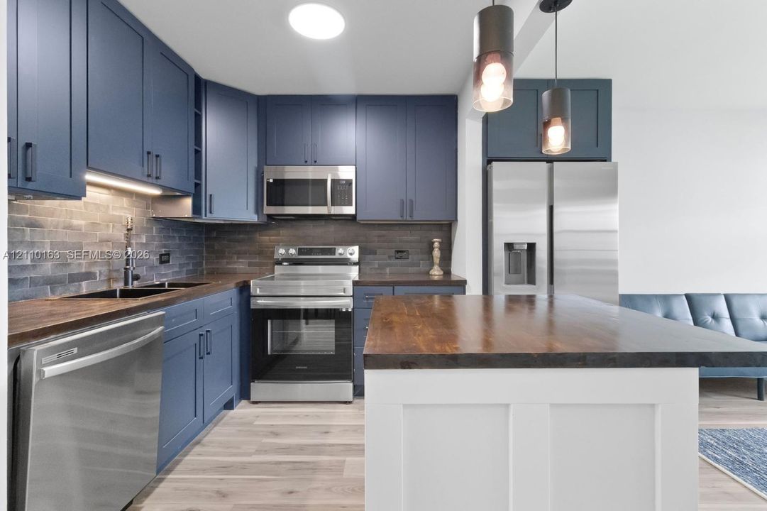 Kitchen featuring navy blue cabinetry, wood-finish countertops, stainless steel appliances, brick-style tile backsplash, and a central island with a white base