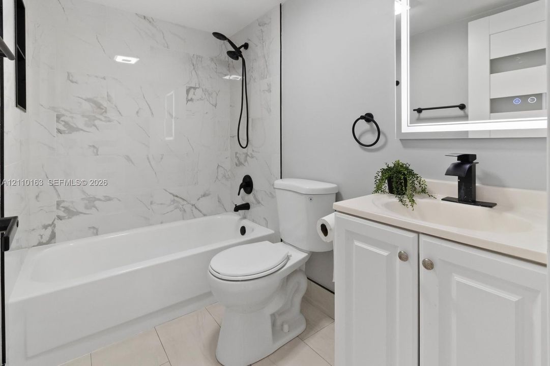Bathroom featuring a soaking tub with full-height stone-look tile surround, vanity with an integrated sink, matte black faucet hardware, and a backlit mirror