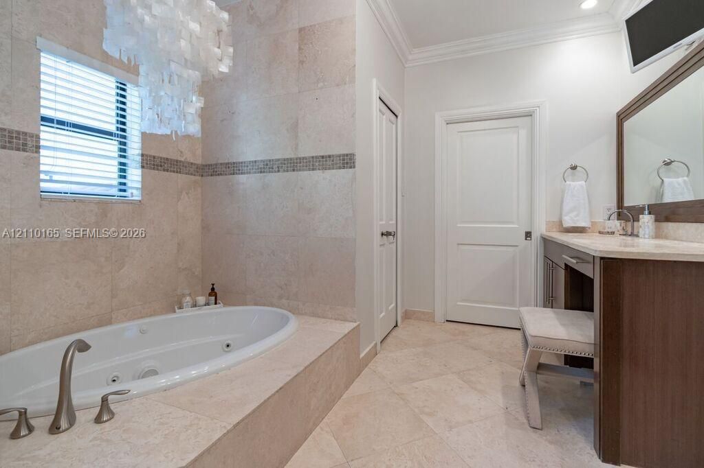 Jetted soaking tub with tile surround, stone-tiled flooring, stone wall accents, vanity with stone countertop, and crown molding