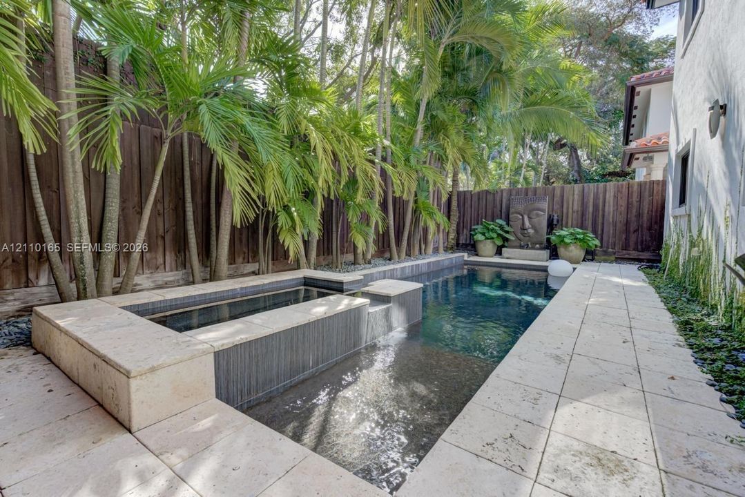 In-ground swimming pool featuring a built-in spa, stone-tile decking, vertical wood fencing, and mature palm trees