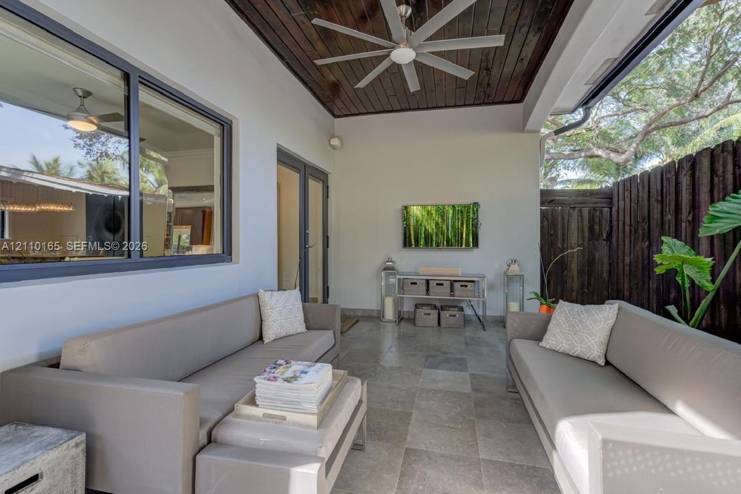 Covered patio featuring large-format stone flooring, wood-paneled ceiling, ceiling-mounted fan, and dark-finished perimeter fencing