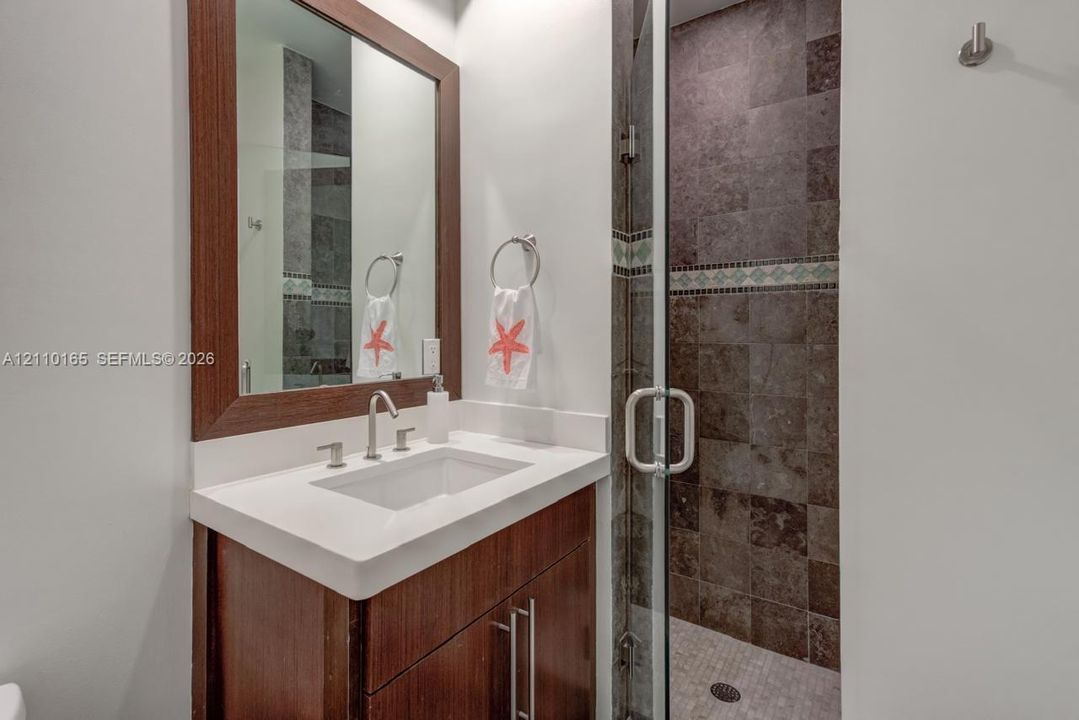 Bathroom vanity with wood-finish cabinetry, white countertop, integrated rectangular sink, framed mirror, and a walk-in shower with stone tile surround, glass enclosure, and pebble-style shower floor