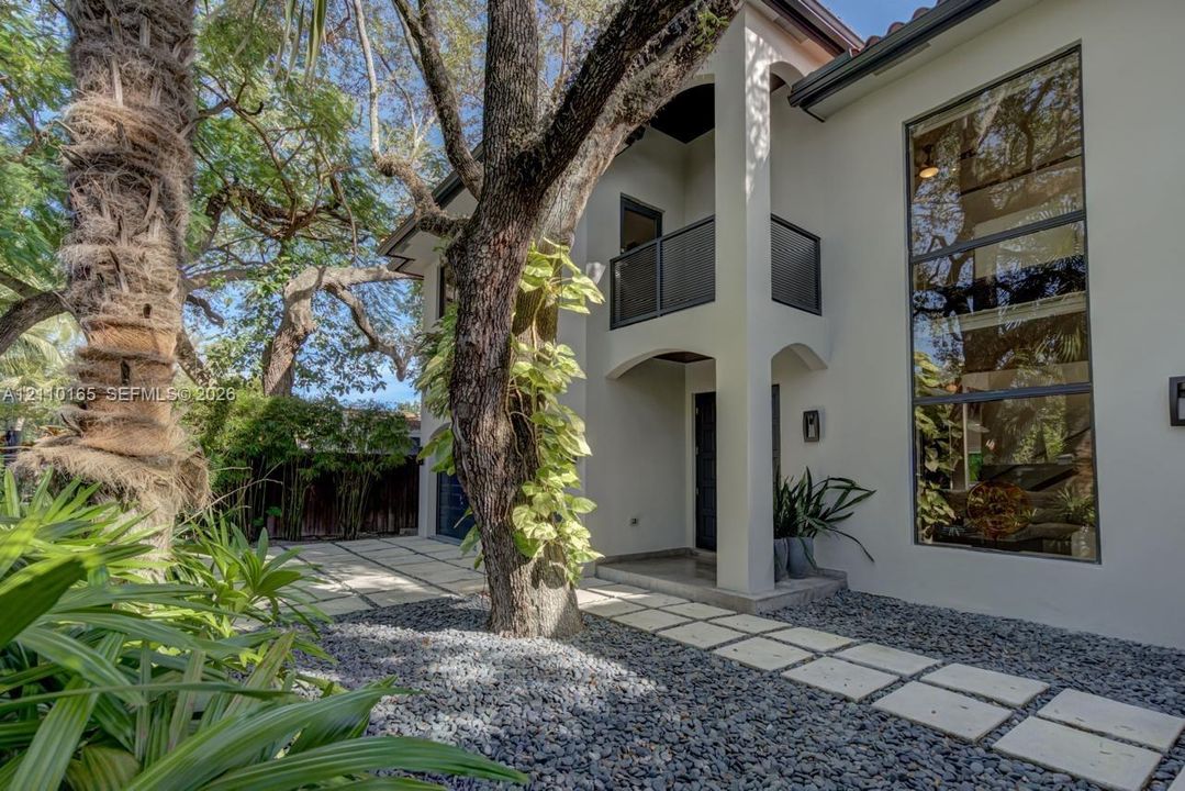 Stucco exterior featuring multi-story floor-to-ceiling windows, a covered entryway with arched supports, a private balcony with metal railing, and a stone paver walkway set in decorative gravel