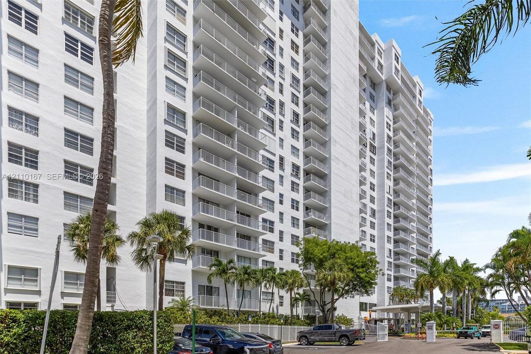 High-rise residential building featuring white stucco facade, stacked private balconies, covered porte-cochère entry, and landscaped tropical grounds with mature palm trees