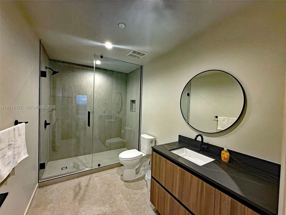 Modern bathroom featuring a wood-finish floating vanity with a dark stone countertop, an integrated rectangular sink, a glass-enclosed walk-in shower with matte black fixtures, and a circular wall-mounted mirror
