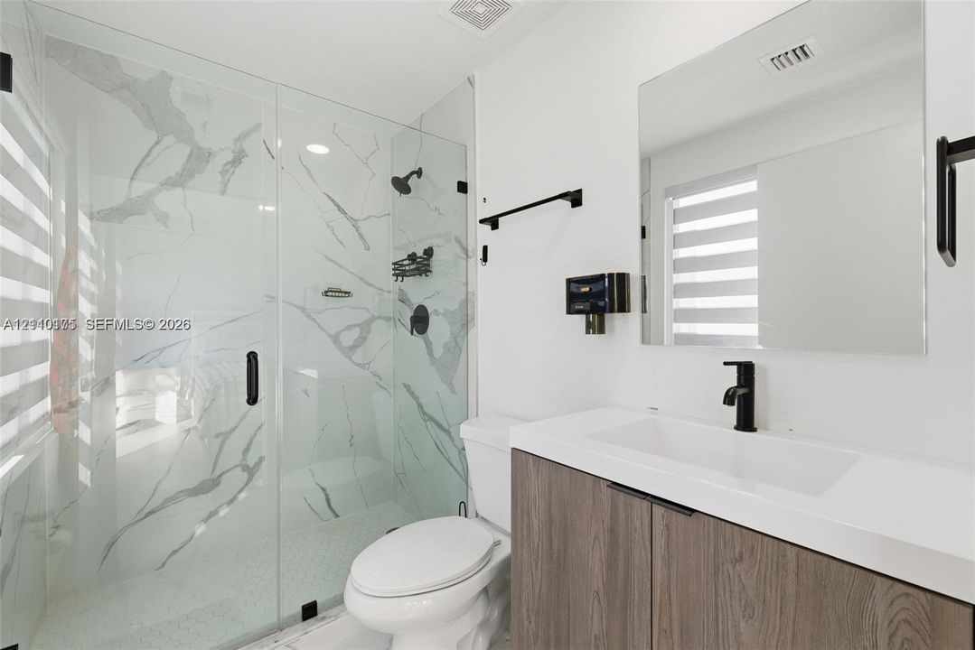 Walk-in shower with stone-finish tile surround and glass enclosure, wood-finish floating vanity with white countertop, matte black fixtures, and large frameless mirror