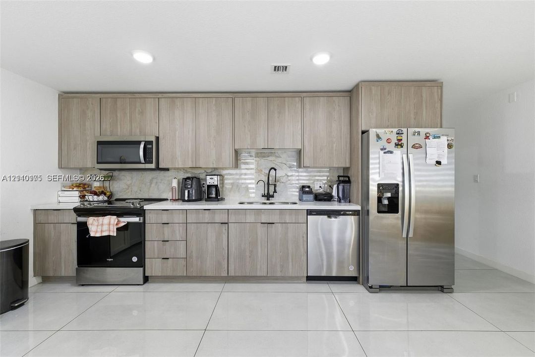 Kitchen featuring wood-finish cabinetry, light-toned stone-style backsplash, integrated stainless steel appliances, and large-format tile flooring