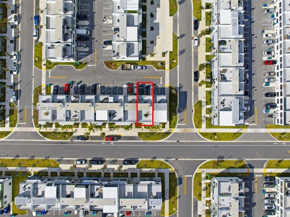 Aerial view of a residential unit featuring a paved rear patio, designated parking spaces, and proximity to internal roadways within the community