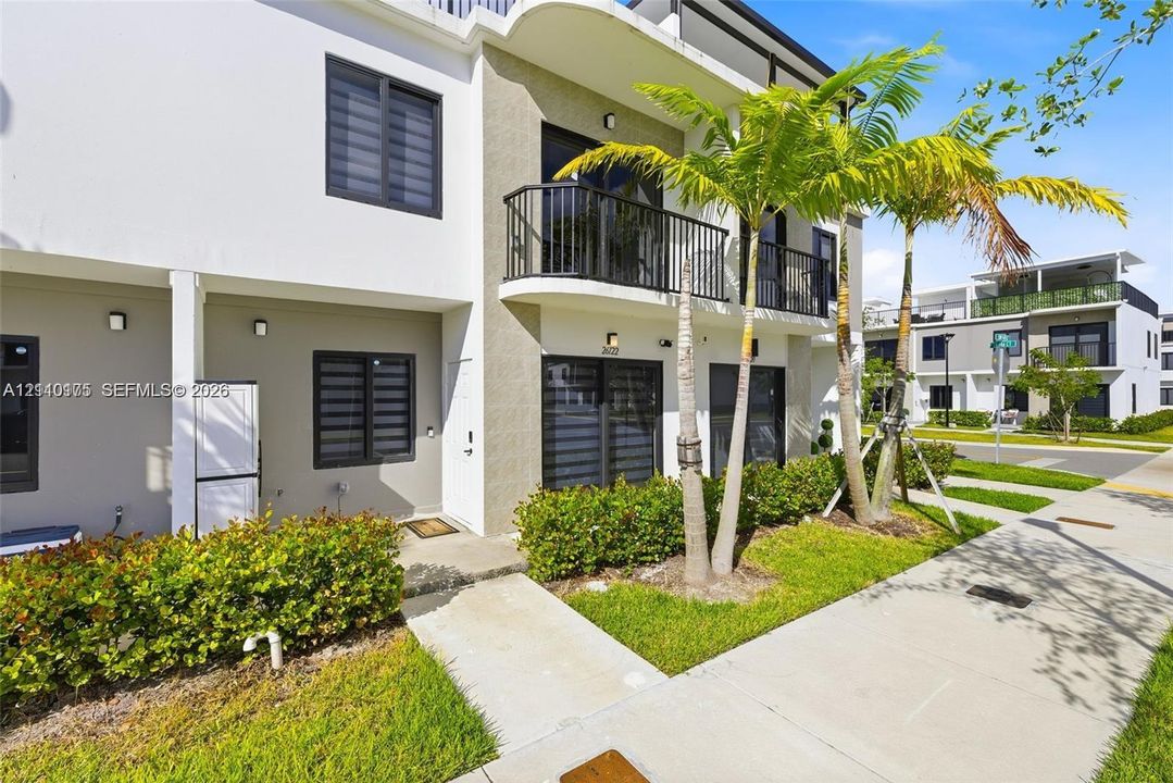 Two-story exterior featuring a stucco facade, second-floor balcony with black metal railing, concrete entry path, and mature palm landscaping