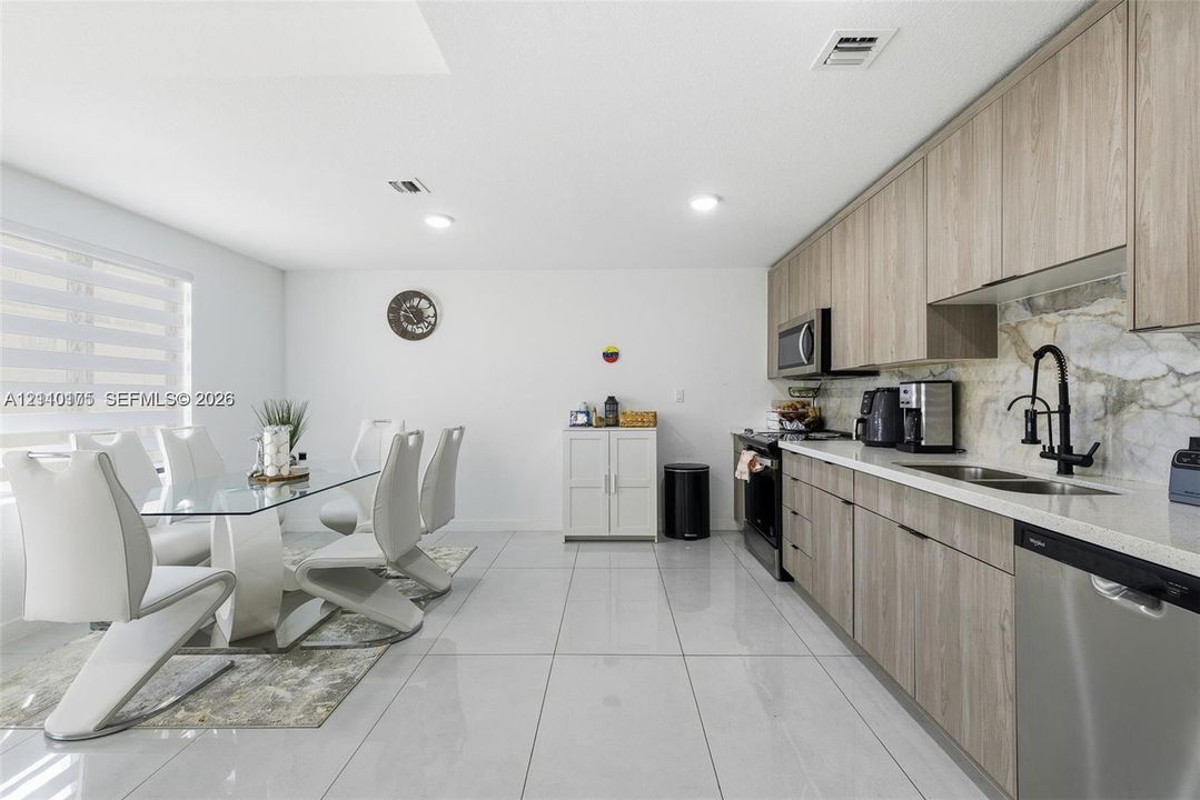 Kitchen featuring wood-finish cabinetry, stone-patterned backsplash, light-colored stone countertops, stainless steel appliances, and large-format porcelain tile flooring