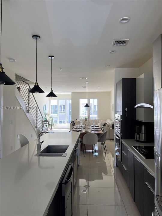 Kitchen featuring a large quartz-topped island with an integrated sink, dark wood-finish cabinetry, built-in stainless steel double oven, polished tile flooring, and decorative pendant lighting