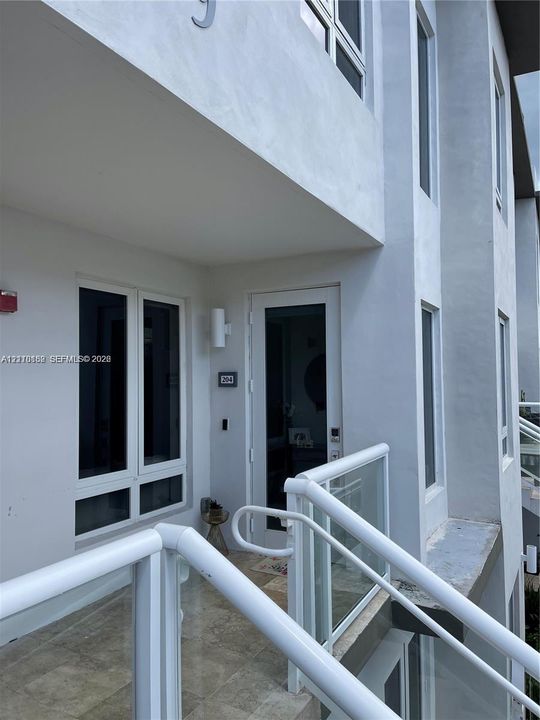 Modern exterior entry featuring a glass-panel balcony railing, tile flooring, vertical multi-pane windows, and a wall-mounted light fixture