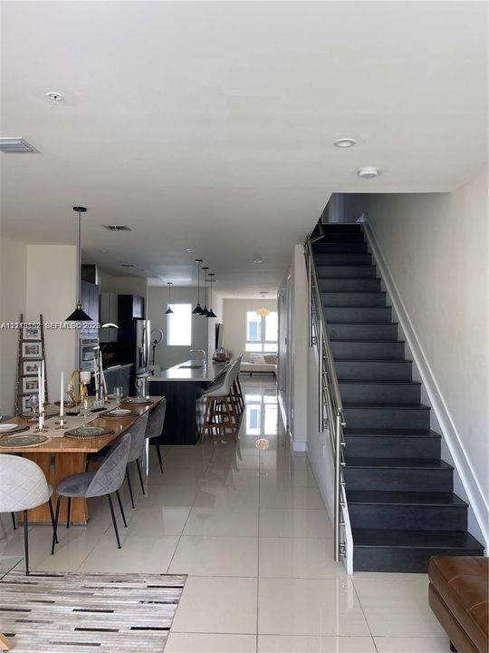 Open-concept interior featuring large-format tile flooring, a kitchen island with stone countertop, stainless steel pendant lighting, and a staircase with metal railings