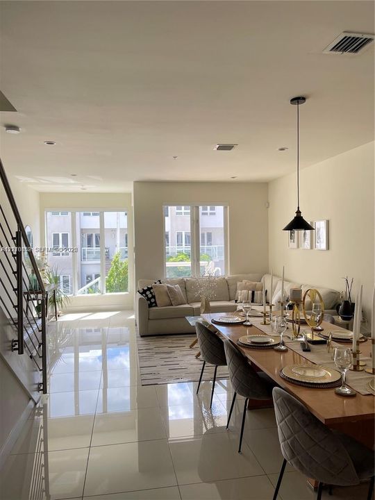 Open-concept interior featuring large-format tile flooring, oversized windows, metal-railed staircase, and a suspended cone-shaped pendant light