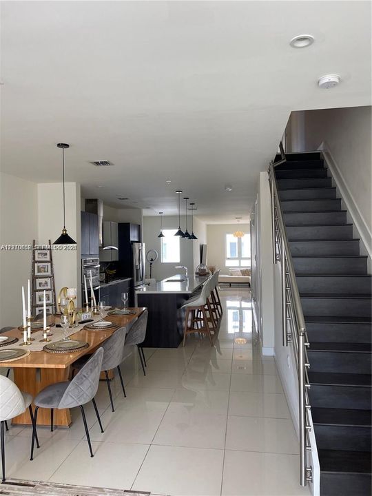Open-concept interior featuring large-format tile flooring, a kitchen island with stone countertops, recessed lighting, and a staircase with a stainless steel railing