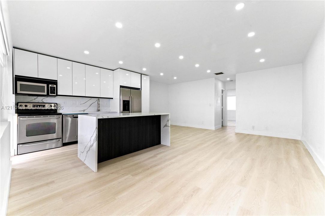 Kitchen featuring high-gloss white cabinetry, a center island with a stone-finish waterfall countertop, stainless steel appliances, and recessed lighting over wood-finish flooring