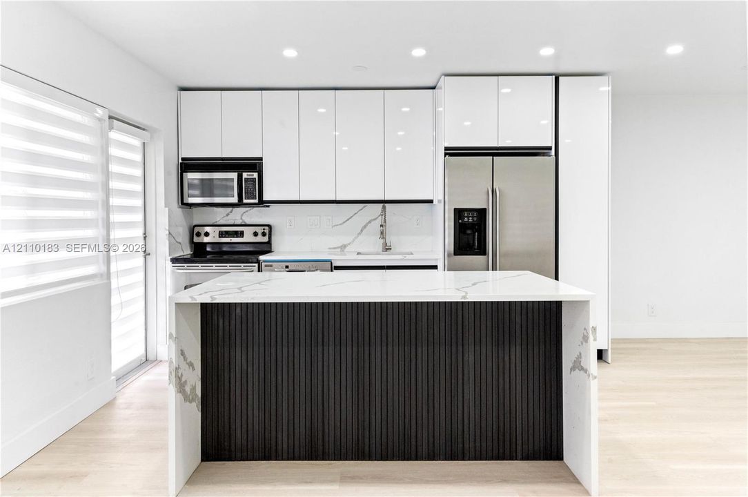 Modern kitchen featuring high-gloss white cabinetry, an island with a waterfall-edge countertop and dark vertical-slat paneling, stainless steel appliances, and light-toned wood-finish flooring