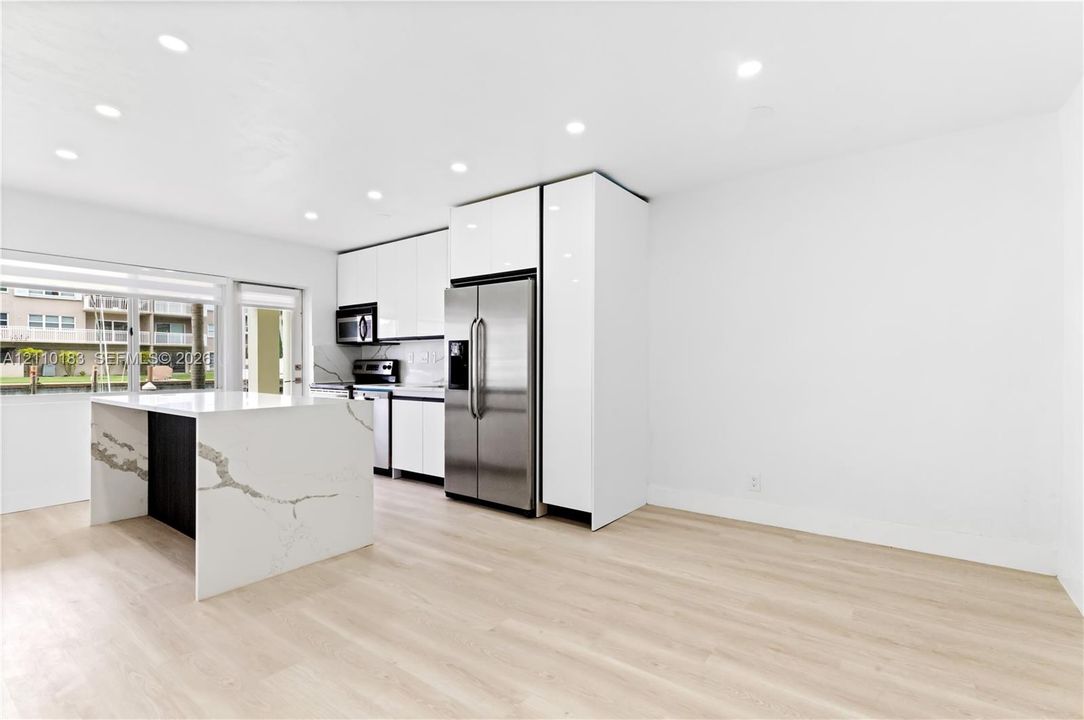 Kitchen featuring high-gloss white cabinetry, a waterfall-edge island with a stone-finish countertop, stainless steel refrigerator, and wood-finish flooring