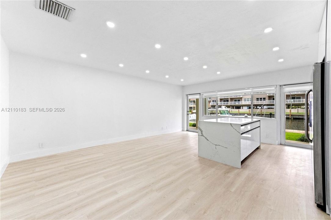Open-concept interior featuring light wood-finish flooring, recessed lighting, and a kitchen island with a white stone-surface countertop and waterfall edge