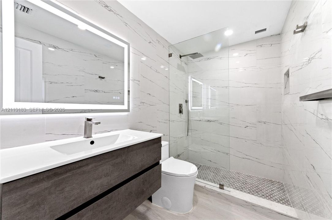 Floating vanity with integrated sink, backlit LED mirror, walk-in shower with frameless glass enclosure, floor-to-ceiling stone-style wall tiling, and mosaic tile shower flooring