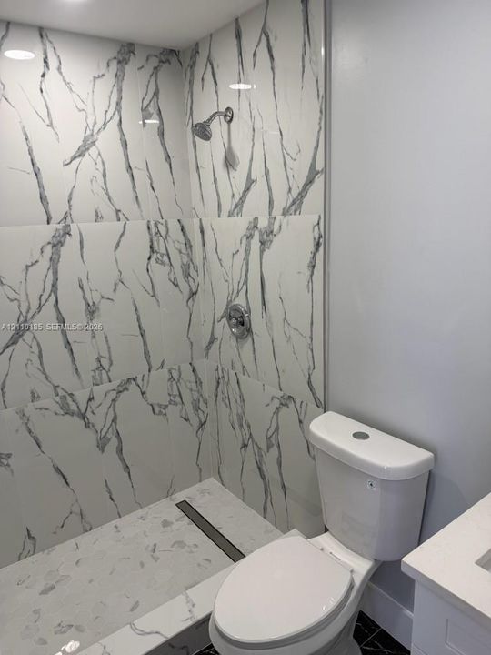 Shower enclosure featuring large-format veined tile surround, chrome shower head, integrated linear drain, and a white finish toilet
