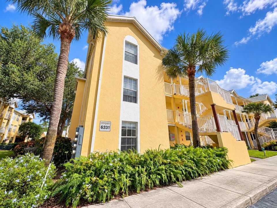 Multi-story exterior featuring a yellow stucco facade, white trim accents, arched window detailing, tiered exterior staircases with white railings, and mature palm tree landscaping